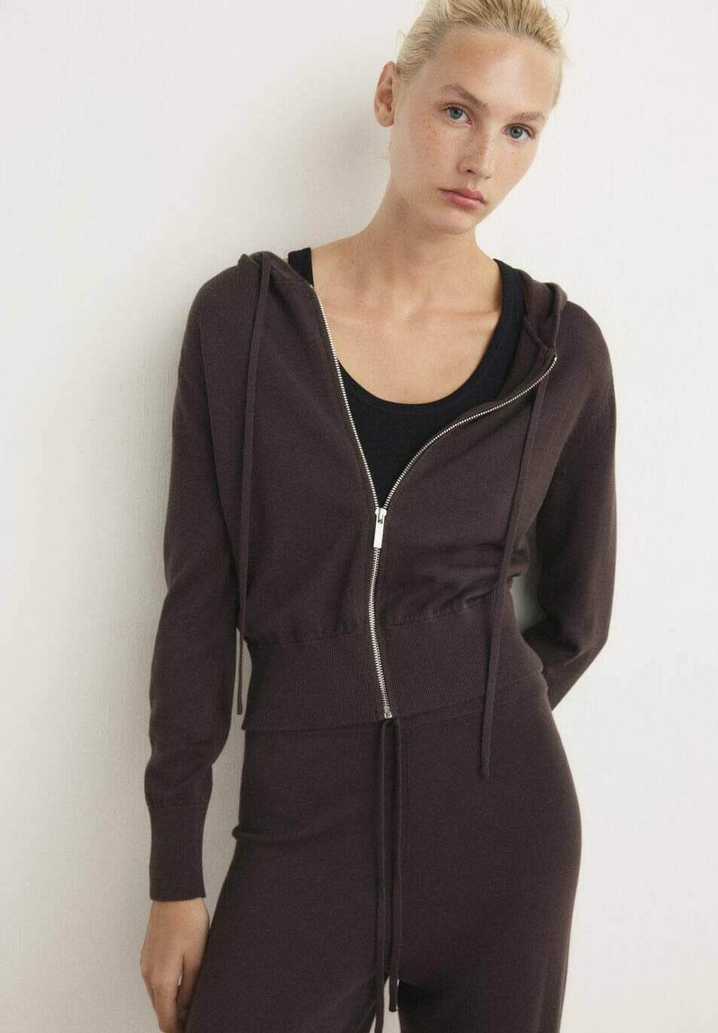 Felpa corta marrone con zip realizzata in tessuto morbido, dotata di cordino, abbinata a un top nero attillato. Design semplice, senza pattern o decorazioni.