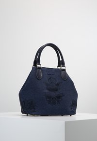 Sac à main bleu foncé avec deux anses, broderie florale subtile et forme trapézoïdale légèrement arrondie sur une surface blanche.
