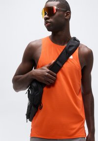 Αθλητικός άνδρας με πορτοκαλί αμάνικη μπλούζα, ανακλαστικά γυαλιά ηλίου και μαύρη τσάντα χιαστί, κοιτάζει προς τα πλάγια με φόντο λευκό.