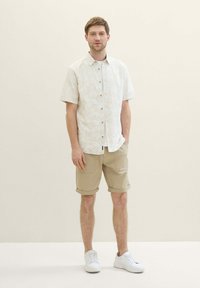 Kurzarmhemd in hellbeige mit Palmblattmuster, kombiniert mit hellbeigen, hochgerollten Shorts und weißen Sneakern.