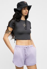 Γυναίκα που φοράει μαύρο καπέλο τύπου bucket Santa Cruz, σκούρο γκρι κοντό μπλουζάκι με κοπή σε σχήμα λουλουδιού και λεβαντίνι σορτς με κορδόνι, με τα χέρια στις τσέπες.