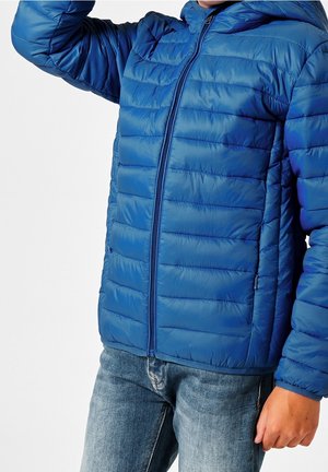 Barn iført en blå quiltet jakke med lynlås og denim jeans, arm løftet, mod en ensfarvet hvid baggrund.