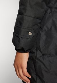 Černý prošívaný rukáv bundy s žebrovaným lemem a knoflíkovým detailem. Tkanina má hladkou strukturu s viditelnými švy.