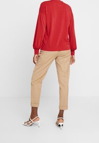Maglietta rossa a maniche lunghe con polsini arricciati, abbinata a pantaloni beige su misura e sandali bianchi con tacco. Tessuto morbido, vestibilità comoda.