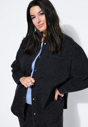 Ung kvinde med langt mørkt hår iført en sort broderet knap-op jakke over en lyseblå top, smilende med den ene hånd på hoften.