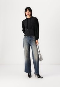 Sudadera negra con capucha y cordones, combinada con jeans de pierna ancha en un tono azul claro. El modelo lleva un bolso plateado y botas de tacón negras.