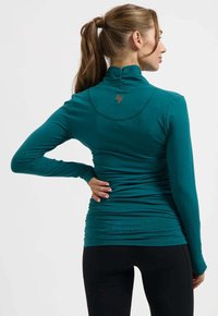 Kvinna med brun hästsvans som bär en turkos långärmad topp och svarta byxor, står med en hand på höften och vänder sig mot höger.