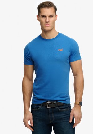 Mand iført blå kortærmet T-shirt med lille orange logo, mørke jeans, sort bælte og armbåndsur, står foran hvid baggrund.