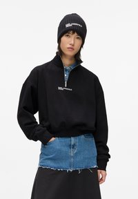 Sudadera negra con cremallera y cuello alto, con el logo en blanco "KARL LAGERFELD JEANS", superpuesta sobre una falda de mezclilla azul, combinada con un gorro de punto negro.