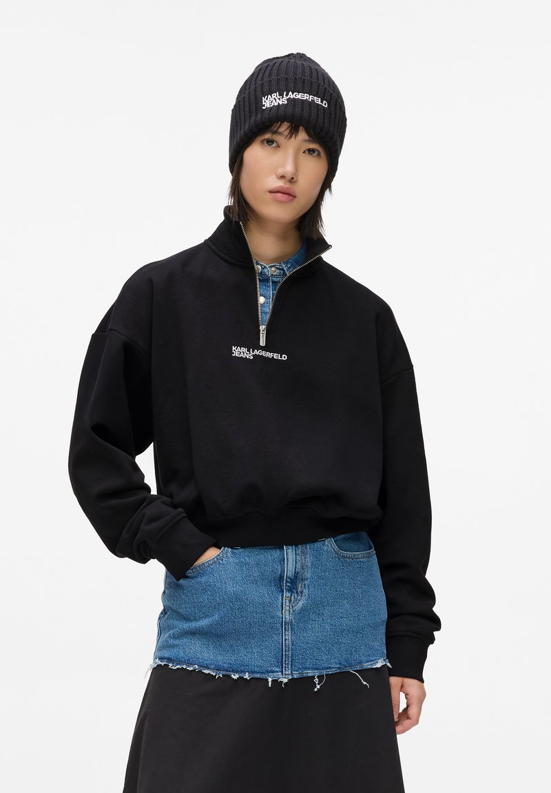 Sudadera negra con cremallera y cuello alto, con el logo en blanco "KARL LAGERFELD JEANS", superpuesta sobre una falda de mezclilla azul, combinada con un gorro de punto negro.