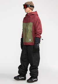 Röd och olivgrön jacka med en stjärnlogotyp, svarta handskar, lösa svarta byxor och svarta sneakers med vita accenter, lämplig för kallt väder.