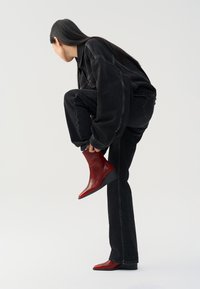 Donna che indossa un completo di jeans neri, mentre si piega per mettere stivali alla caviglia in pelle rossa lucida con tacchi a zeppa, su uno sfondo semplice.