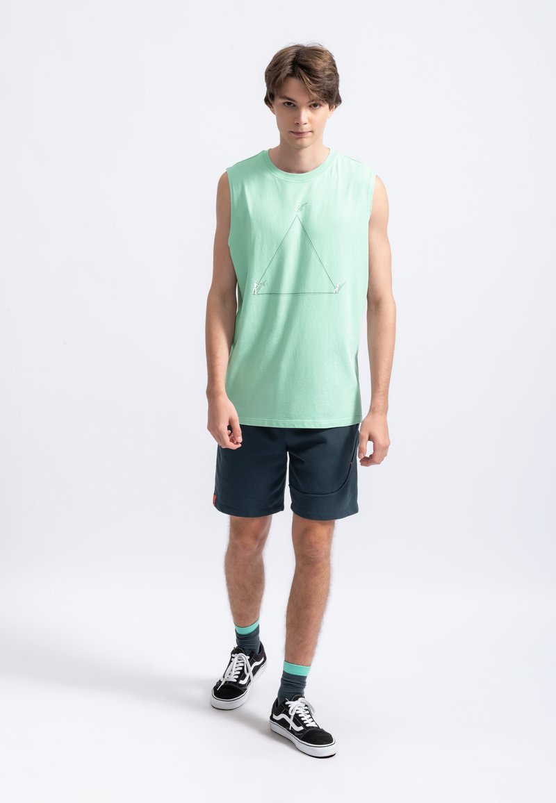 Maglietta senza maniche verde menta con una grafica triangolare bianca geometrica, abbinata a pantaloni corti blu navy e scarpe da ginnastica nere.