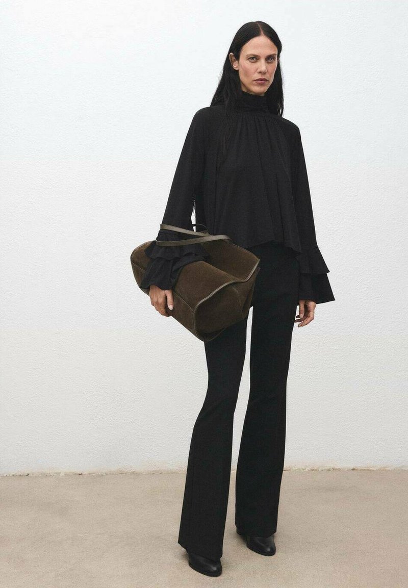 Top negro de manga larga con cuello alto y puños con volantes, combinado con pantalones negros ajustados y un bolso de ante marrón. Texturas suaves, líneas limpias.