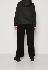 Persona vestida con chaqueta negra con capucha, pantalones negros de pierna ancha y zapatos negros de suela gruesa, de pie frente a un fondo claro y liso.