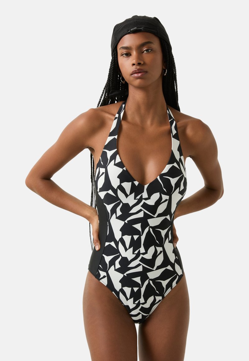 Maillot de bain une pièce noir et blanc à motif avec un décolleté en V profond et des bretelles halter. Le design présente des formes abstraites et courbées ainsi qu'un tissu texturé.