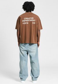 Brown cotton t-shirt with large white text "CREATIVE DEPARTMENT 2018 CD" on the back, paired with light blue baggy jeans and white shoes.