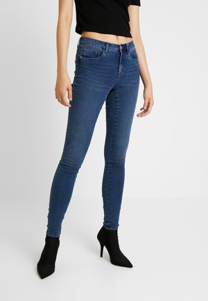 Femme portant un jean skinny taille mi-haute bleu, un top court noir et des bottines noires à talons hauts et bouts pointus, debout devant un fond uni.