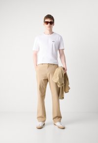 Tricou alb cu maneci scurte și logo negru, pantalonii bej cu croi drept, jachetă bej purtată pe braț, adidași albi cu talpă bej, ochelari de soare maroon.