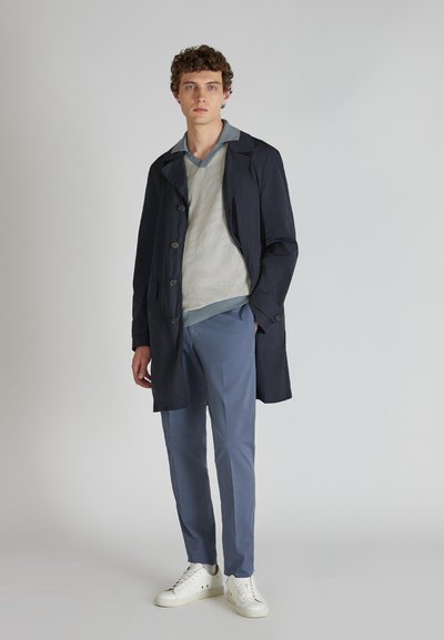 Marineblauer leichter Trenchcoat mit zwei Knöpfen, über einem cremefarbenen strukturierten Pullover und hellblauen Hosen getragen, kombiniert mit weißen Sneakers.