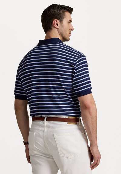 Tricou polo cu dungi bleumarin și alb, cu mâneci scurte și guler, asortat cu pantaloni albi și o curea maro. Textură de material neted.