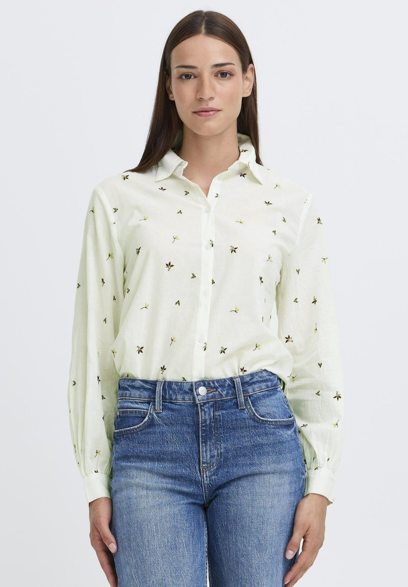 Lys grøn knap-op skjorte med blomsterbroderi, parret med blå denimjeans. Skjorten har en krave og lange ærmer.