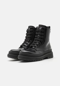 Bottines en cuir verni noir avec lacets plats noirs, semelles épaisses texturées et détails de couture surélevés sur les côtés et autour des orteils.
