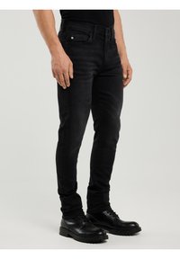 Bărbat purtând blugi negri slim-fit și pantofi negri din piele cu șireturi, stând în picioare pe un fundal simplu, deschis la culoare.