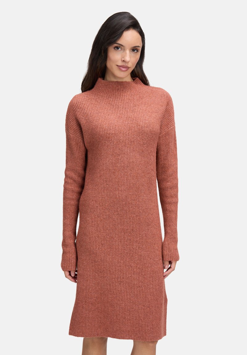 Vrouw met lang donker haar, gekleed in een geribbelde, lange mouw, knielange trui-jurk in roestkleur, staand tegen een effen achtergrond.