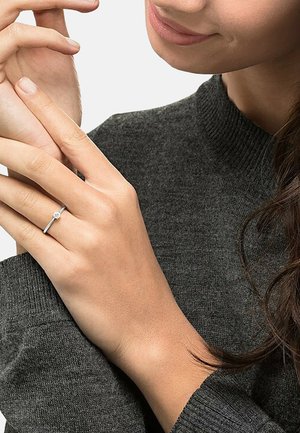 Silberner Ring mit einem runden Stein, getragen an einem schlanken Finger, vor einem dunkelschwarzen Strickpullover-Hintergrund. Nahaufnahme.