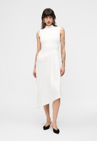 Γυναίκα με τατουάζ που φοράει λευκό midi φόρεμα χωρίς μανίκια και μαύρα μυτερά ψηλοτάκουνα, στέκεται μπροστά σε απλό ανοιχτό φόντο.