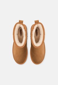 Bottines en daim marron avec doublure en tissu crème pelucheux, bouts ronds et languettes en textile pour un enfilage facile. Extérieur lisse avec une texture douce.