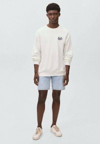 Sweatshirt crème avec un petit logo, associé à un short en denim clair. La tenue est complétée par des baskets beiges, sur un fond uni.