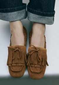 Brown suede moccasin-style shoes with fringed detailing and a tied bow, paired with rolled-up dark denim pants.