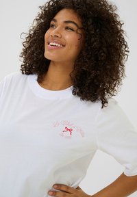 T-shirt en coton blanc à manches courtes, col rond, avec un logo brodé rouge représentant un nœud et du texte. Texture lisse et coupe décontractée.