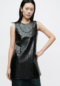 Svart ermeløs topp i kunstskinn med en glatt tekstur, med et halskjede-lignende svart pynteelement ved halsen og avslappet passform.