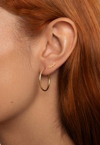 Pendiente de aro dorado con un acabado suave junto a tres pequeños pendientes en forma de estrella dorados; todo ello sobre un fondo de cabello rojo.