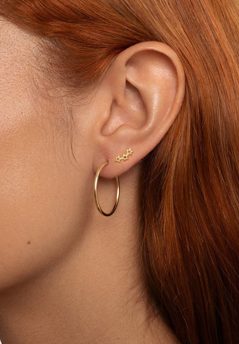 Pendiente de aro dorado con un acabado suave junto a tres pequeños pendientes en forma de estrella dorados; todo ello sobre un fondo de cabello rojo.