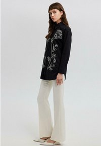 Black button-up shirt with white floral embroidery, paired with fitted cream trousers and light shoes. Minimalist design and sleek silhouette.