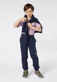 Kind dat marineblauwe joggingbroek en mauve t-shirt draagt, heeft een marineblauwe sweatshirt om zijn schouders gebonden en staat op een witte achtergrond.