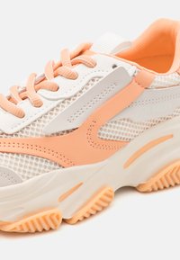 Zapatilla blanca y melocotón con paneles de malla, punta redonda y suela texturizada. Cuenta con cordones rosas, un cuello acolchado y una marca en el costado.