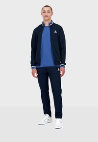 Giacca navy con zip e dettagli bianchi, t-shirt blu, pantaloni navy e sneakers bianche. Presenta il logo sul petto e sui pantaloni.