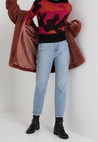 Persona che indossa un cappotto di pelliccia marrone aperto su un maglione a fantasia rosso, nero e rosa, jeans azzurri chiari e stivali alla caviglia neri con borchie.