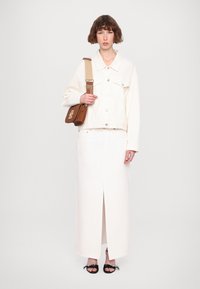 Giacca di denim bianco con tasche frontali abbinata a una gonna maxi bianca con uno spacco frontale. Borsa a spalla marrone e sandali neri.