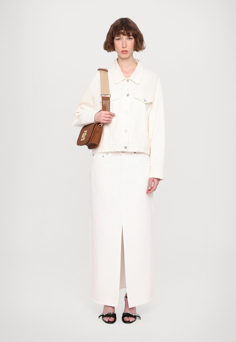 Giacca di denim bianco con tasche frontali abbinata a una gonna maxi bianca con uno spacco frontale. Borsa a spalla marrone e sandali neri.