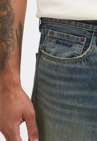Jeans de ganga em azul desbotado com uma ligeira textura. Apresenta um bolso frontal com um logótipo visível, rebites em cobre e uma borda desgastada.