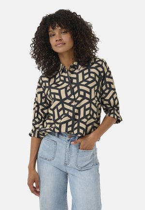 Chemise boutonnée crème avec un bold motif géométrique noir. Présente des manches retroussées et une coupe décontractée. Associée à un jean en denim clair.
