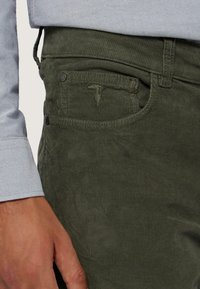 Zielone spodnie sztruksowe o prążkowanej fakturze, z przednią kieszenią i kontrastowym przeszyciem. Ręka znajduje się na pasku.