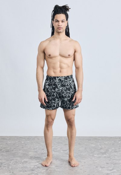 Άνδρας χωρίς πουκάμισο με μακριά dreadlocks, ξυπόλυτος, στέκεται σε πέτρινο δάπεδο φορώντας μαύρα και λευκά μαγιό με σχέδια.