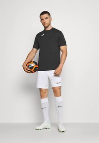 Férfi fekete sportpólóban, fehér rövidnadrágban, zoknikban és stoplis cipőben, egy fekete és narancssárga focilabdát tart a sima háttér előtt.